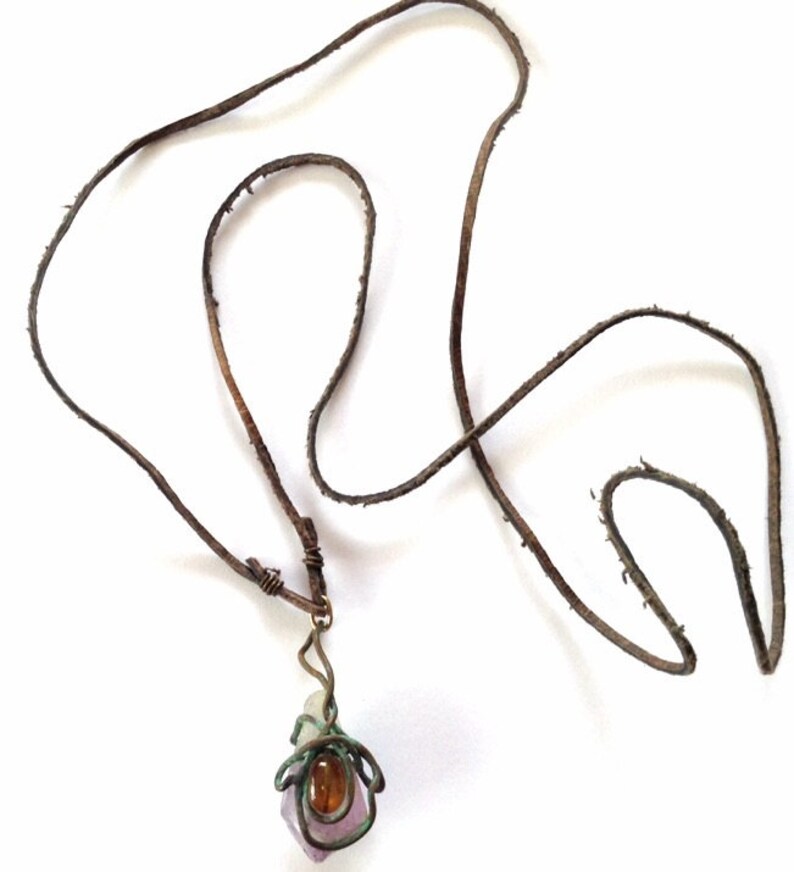 May include: A brown leather cord necklace with a pendant featuring a purple and clear crystal wrapped in copper wire. A small orange gemstone is visible in the center of the pendant.