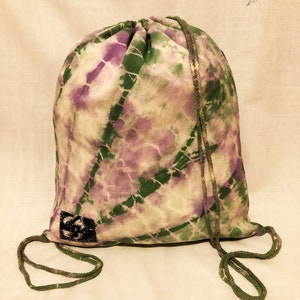 May include: A green and purple tie-dye drawstring bag with a black square label. The bag is made of a soft, lightweight fabric and has a simple design.