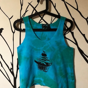 May include: A blue and green tie-dye tank top with a black cupcake graphic. The tank top has a scoop neck and a relaxed fit.