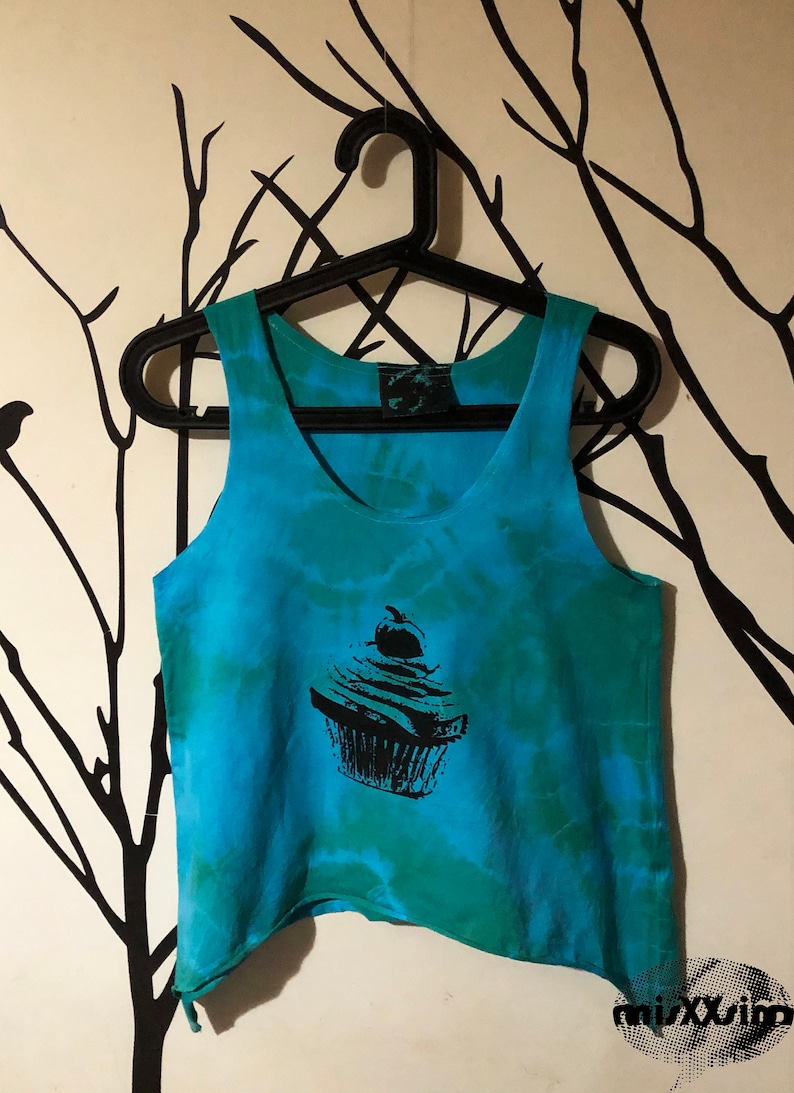 May include: A blue and green tie-dye tank top with a black cupcake graphic. The tank top has a scoop neck and a relaxed fit.