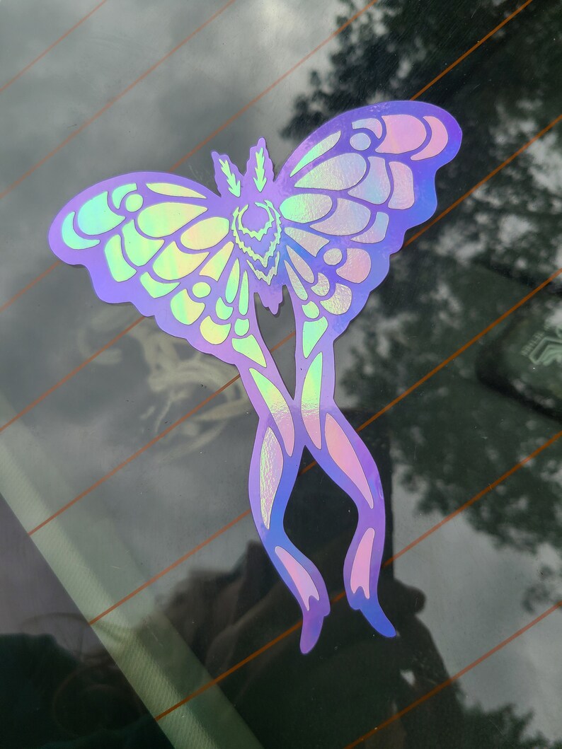 May include: A holographic iridescent sticker of a moth with wings spread out. The moth is a light purple color with a green and blue iridescent sheen.
