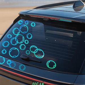 Puede incluir: La ventana trasera de un coche gris oscuro está decorada con calcomanías en forma de burbujas de color turquesa. La palabra "OCEAN" se muestra en texto verde en la parte trasera del coche. El fondo muestra una playa y el océano.
