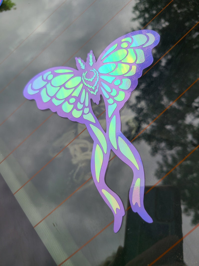 May include: A holographic iridescent sticker of a moth with a green and purple color scheme. The moth has a large wingspan and intricate details.
