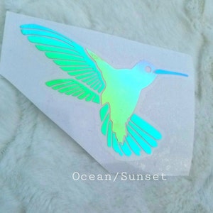 May include: A iridescent green and blue hummingbird decal with a white background. The decal has the text "Ocean/Sunset" printed below the hummingbird.