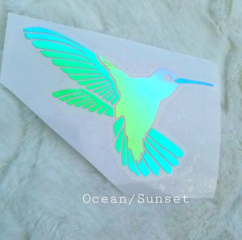 May include: A iridescent green and blue hummingbird decal with a white background. The decal has the text "Ocean/Sunset" printed below the hummingbird.
