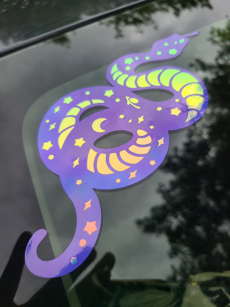 Pode incluir: Um adesivo de vinil iridescente roxo e verde de uma cobra com estrelas e luas no corpo.