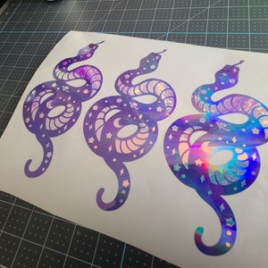 Pode incluir: Tr&ecirc;s adesivos de cobra roxo iridescentes com um design celestial. As cobras est&atilde;o enroladas e t&ecirc;m estrelas e luas em seus corpos.