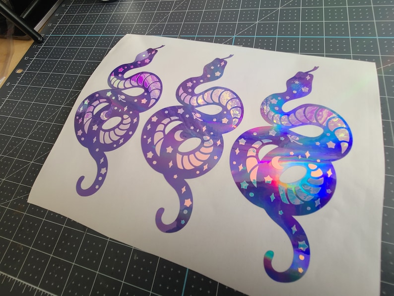 Pode incluir: Tr&ecirc;s adesivos de cobra roxo iridescentes com um design celestial. As cobras est&atilde;o enroladas e t&ecirc;m estrelas e luas em seus corpos.