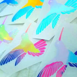 May include: A set of iridescent hummingbird stickers in various colors, including blue, yellow, green, pink, and purple. The stickers are on white paper.