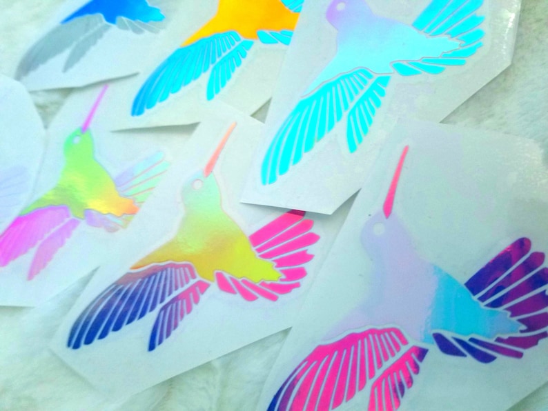 May include: A set of iridescent hummingbird stickers in various colors, including blue, yellow, green, pink, and purple. The stickers are on white paper.