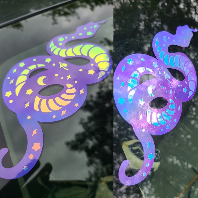 Pode incluir: Dois adesivos de vinil hologr&aacute;ficos iridescentes de uma cobra com estrelas e luas. A cobra est&aacute; enroscada em um c&iacute;rculo com a cauda na boca. Os adesivos est&atilde;o na janela de um carro.