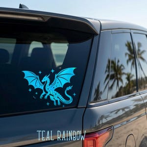 May include: A teal dragon sticker with outstretched wings and star accents is affixed to a car window. The sticker has a gradient effect, transitioning from a darker teal to a lighter shade. The text "TEAL RAINBOW" is displayed below the dragon.