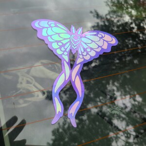 May include: A holographic sticker of a purple and green butterfly with intricate details. The butterfly is on a car window.