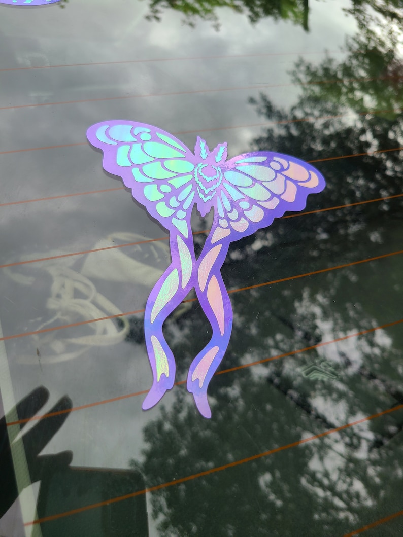 May include: A holographic sticker of a purple and green butterfly with intricate details. The butterfly is on a car window.