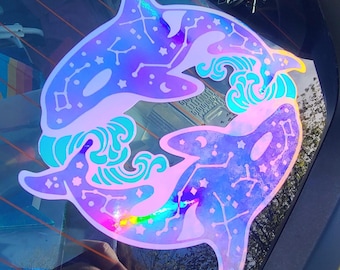 May include: A holographic sticker featuring two orcas in a circular design. The orcas are purple and blue with constellations and stars. Turquoise waves and accents surround the orcas. The sticker is displayed on a car window.