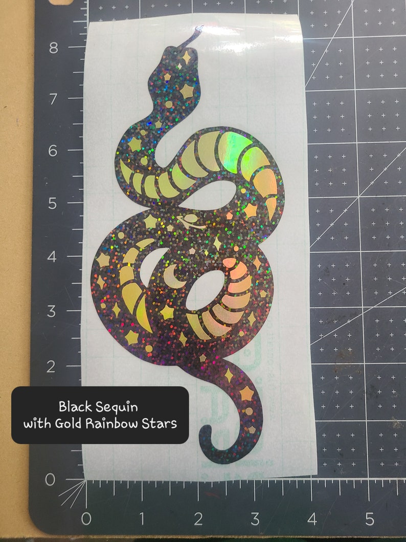 Pode incluir: Cobra de lantejoulas pretas com estrelas arco-&iacute;ris douradas. A cobra est&aacute; enroscada em um c&iacute;rculo com a cauda na boca.