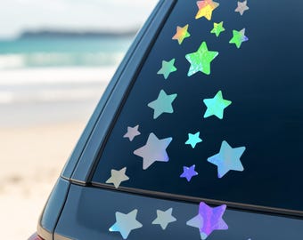 May include: A collection of iridescent star-shaped decals applied to the rear window of a gray vehicle. The stars vary in size and color, creating a whimsical design. The background shows a beach scene with a blue sky and ocean.