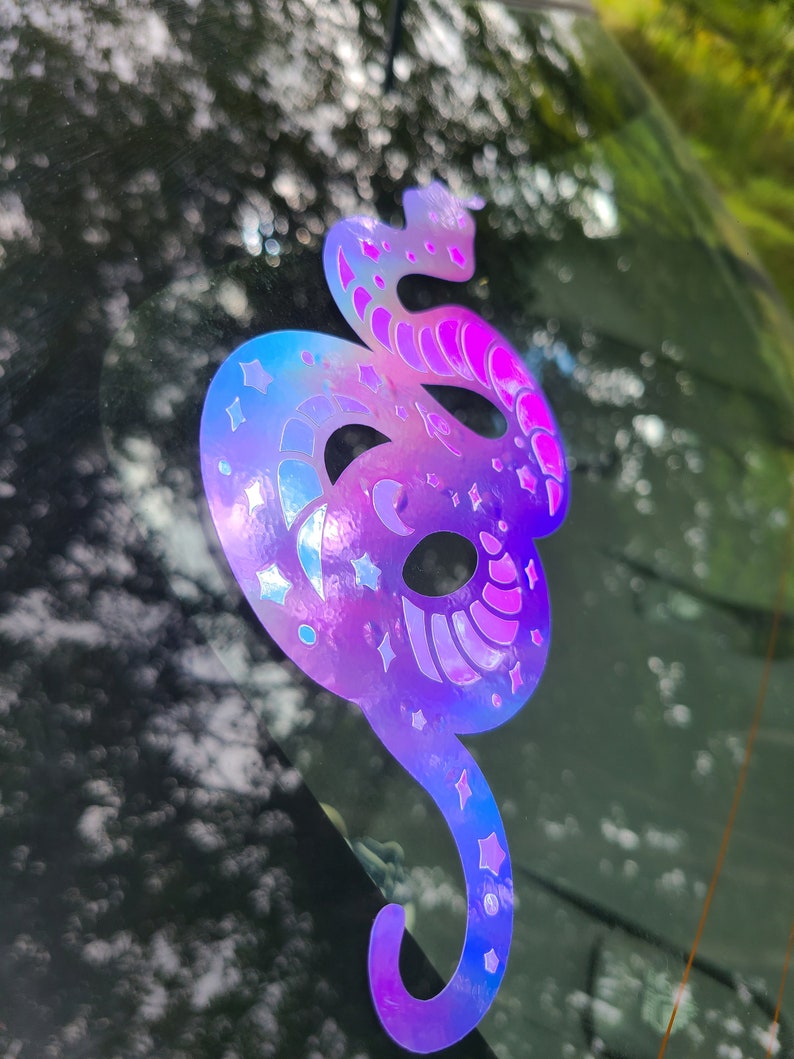 Pode incluir: Um adesivo hologr&aacute;fico iridescente de uma cobra com estrelas e luas no corpo. O adesivo est&aacute; na janela de um carro.