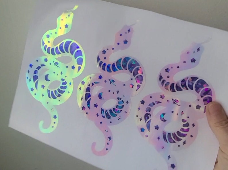 Pode incluir: Tr&ecirc;s adesivos de vinil hologr&aacute;fico iridiscente de uma cobra com estrelas e luas. As cobras s&atilde;o de cores diferentes, uma &eacute; azul claro, outra rosa e outra roxa.