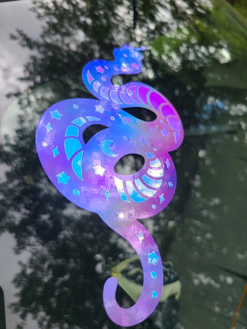 Pode incluir: Um adesivo hologr&aacute;fico de uma cobra com um design celestial. A cobra est&aacute; enroscada e tem um degrad&ecirc; de cores roxo, azul e rosa. O design inclui estrelas e luas crescentes.