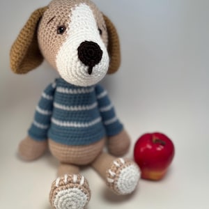 May include: A handmade crochet dog doll with a white and tan head, brown ears, and a blue and white striped shirt. The dog has tan legs and feet with white accents. A red apple sits beside the dog on a white surface.