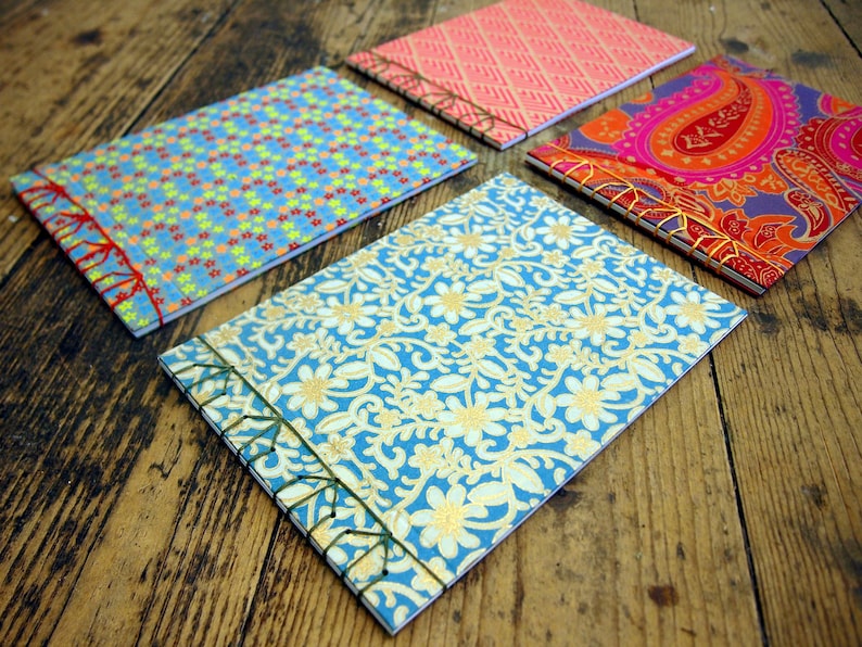 Peut inclure: Quatre petits carnets faits &agrave; la main avec des couvertures &agrave; motifs diff&eacute;rents. Les couvertures sont bleues avec des &eacute;toiles blanches, bleues avec des motifs floraux dor&eacute;s, roses avec un motif g&eacute;om&eacute;trique et violettes avec un motif paisley.