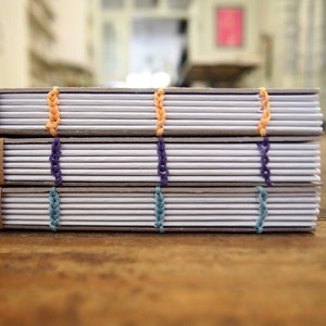Peut inclure: Trois cahiers avec des couvertures brunes et des pages blanches sont empil&eacute;s sur une surface en bois. Les cahiers sont reli&eacute;s avec des coutures color&eacute;es, y compris du fil orange, violet et bleu.