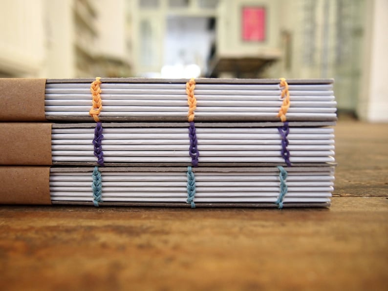 Peut inclure: Trois cahiers avec des couvertures brunes et des pages blanches sont empil&eacute;s sur une surface en bois. Les cahiers sont reli&eacute;s avec des coutures color&eacute;es, y compris du fil orange, violet et bleu.