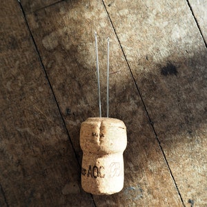 Puede incluir: Un corcho de una botella de vino con dos alambres metálicos que sobresalen de la parte superior. El corcho es marrón y tiene el texto "M.I.A. AOC" impreso en él.