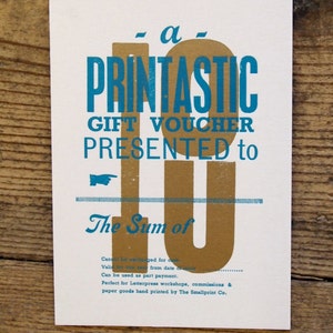 Könnte beinhalten: Ein goldener und blaugrüner Geschenkgutschein mit dem aufgedruckten Text "a- PRINTASTIC GIFT VOUCHER PRESENTED to The Sum of".