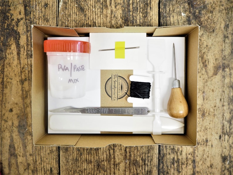 Peut inclure: Une bo&icirc;te en carton marron avec un insert en mousse blanche contenant des outils de travail du cuir. Les outils comprennent un petit pot de colle PVA, une aiguille, une bobine de fil noir, un poin&ccedil;on &agrave; cuir, une r&egrave;gle en m&eacute;tal, un couteau artisanal et une feuille de papier blanche. La bo&icirc;te est &eacute;tiquet&eacute;e "The Smallprint Co."