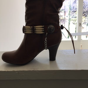 May include: Brown leather knee-high boot with a decorative beaded ankle strap and a small heel. The boot features a zipper and a metal embellishment on the side. The boot is approximately 12 inches tall.