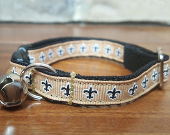 Könnte beinhalten: Ein braunes und schwarzes Katzenhalsband mit einer silbernen Glocke. Das Halsband hat ein schwarzes und weißes Fleur-de-lis-Muster.