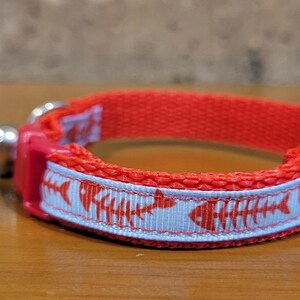 May include: Red cat collar with a light blue band featuring a repeating pattern of red fish skeletons. The collar has a silver bell and a red plastic clasp. This pet accessory is designed for cats.