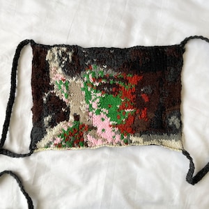 Puede incluir: Una máscara facial de punto con un patrón abstracto colorido en tonos marrones, verdes, rosas y blancos. La máscara tiene lazos negros.