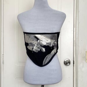 Puede incluir: Un corsé negro y plateado con un patrón geométrico. El corsé está en un maniquí blanco.