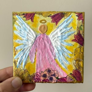 May include: A square painting of an angel with white and light blue wings, a pink robe, and a gold halo. The background is yellow with a floral border in shades of red, pink, and gold. The artwork is held by a hand.