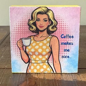 May include: A square canvas print featuring a pop art illustration of a blonde person in a yellow polka dot dress holding a white coffee cup. The background has a pink halftone dot pattern and blue watercolor accents with the text Coffee makes me nice..