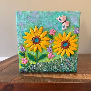 May include: A square, textured artwork featuring two large yellow sunflowers with teal centers, pink and purple flowers, and a butterfly. The background is a mix of green and blue, with a grassy texture at the bottom.