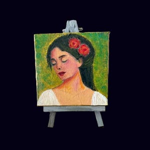May include: A small painting on a miniature easel. The artwork depicts a woman with dark hair adorned with red flowers, set against a green background. The woman has closed eyes and a serene expression. The painting is framed by a white top.