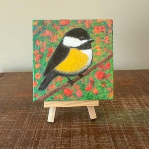 May include: A small, square painting of a bird perched on a branch, displayed on a miniature wooden easel. The bird has a black head, white cheeks, and a yellow breast. The background is a mix of green and red.
