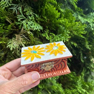 May include: A small, decorative wooden box with a hinged lid. The box is painted with a red and orange pattern on the sides and features two yellow flower designs with blue centers on the top. A small metal clasp secures the lid.