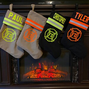 May include: Four personalized Christmas stockings designed to resemble firefighter turnout gear. Two are tan and two are black, each featuring a reflective stripe, a Maltese cross FD emblem, and the names PAUL, CHRISTOPHER, and TYLER in bold lettering.