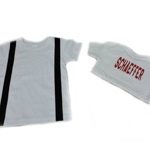 May include: Two white t-shirts. One has two vertical black stripes, resembling suspenders. The other shirt is smaller and has the name "SCHAEFFER" printed in red block letters.