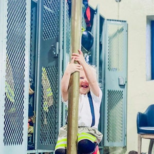 May include: A child in a firefighter's uniform, including red boots, suspenders, and a white shirt, smiles while holding onto a brass pole. The background includes metal lockers and a black base.