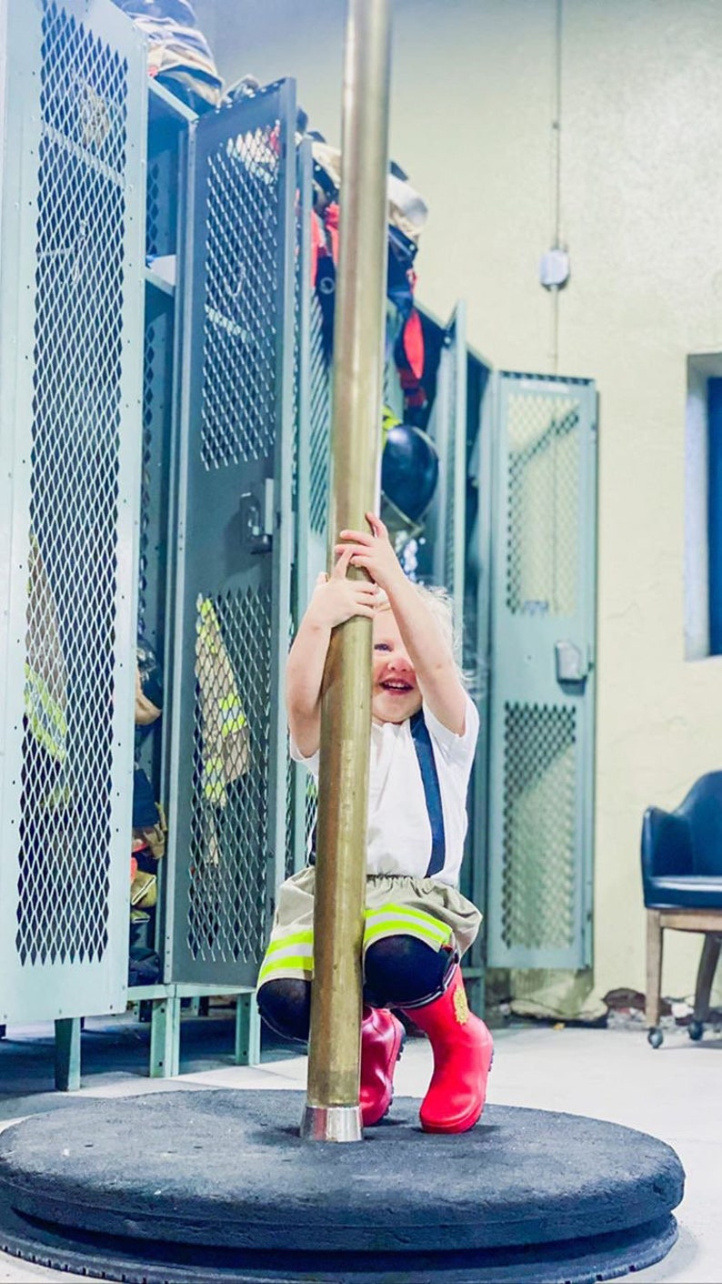 May include: A child in a firefighter's uniform, including red boots, suspenders, and a white shirt, smiles while holding onto a brass pole. The background includes metal lockers and a black base.