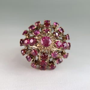 May include: A spherical cluster ring featuring numerous round-cut, deep pinkish-red rubies set in a textured gold metal frame. The design consists of a central ruby surrounded by multiple layers of smaller rubies arranged in a radial, dome-like pattern.