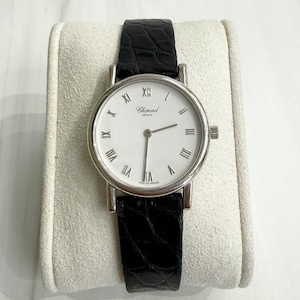 Puede incluir: Un reloj de pulsera Chopard Genève con caja redonda plateada, esfera blanca con números romanos negros y correa de cuero negro texturizado. La esfera muestra el logotipo de Chopard Genève y el texto SWISS MADE en la parte inferior.