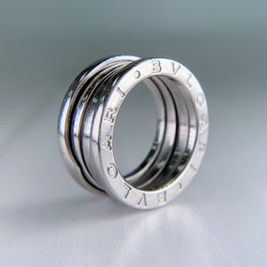 May include: A silver ring with a multi-band design. The ring features the engraved word "BVLGARI" on the outer band. The ring is made of a shiny, reflective metal and is set against a soft, blurred background.