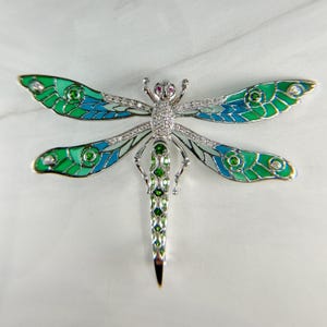 May include: A dragonfly brooch with a silver-tone metal body, featuring green and blue enamel wings. The wings are accented with clear crystals and green gemstones. The dragonfly's head has two red gemstones.
